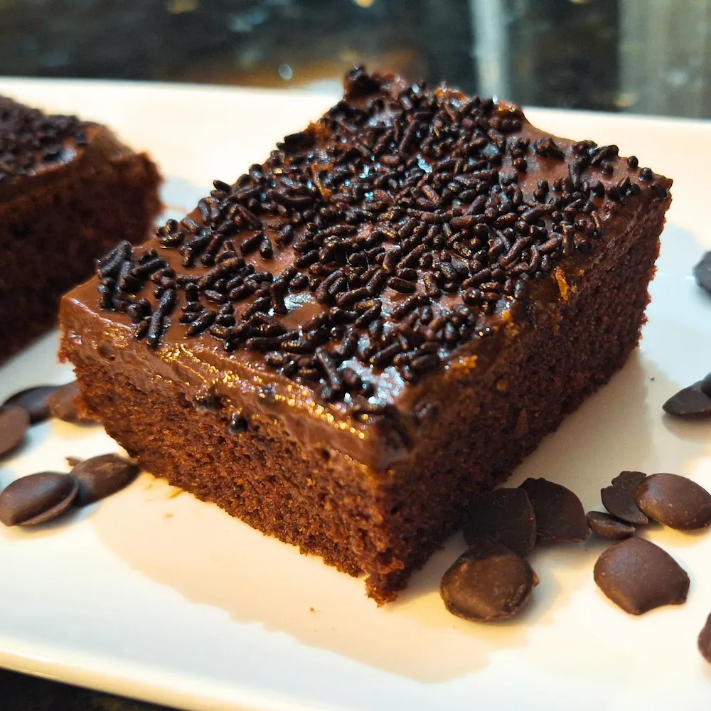 A close-up of a square slice of authentic Moroccan chocolate cake, featuring a moist crumb topped with a rich dark chocolate glaze and sprinkles, served on a white plate.