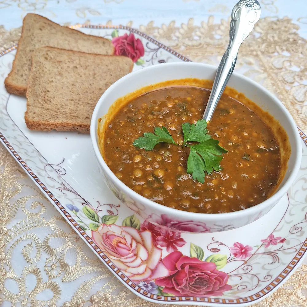 Close-up of a bowl of One-Pot Moroccan Lentils showing the thick, velvety texture and fresh herb garnish next to crusty bread.