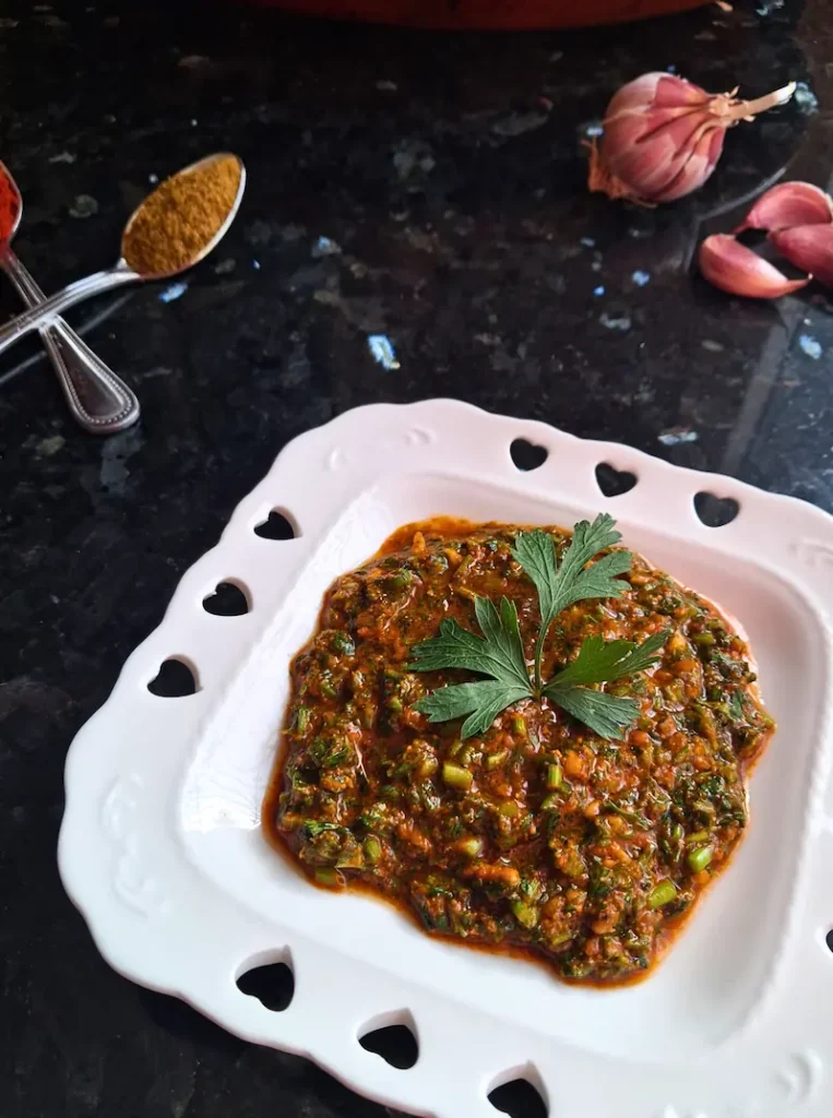 Chermoula_Vertical A close-up, vertical photograph of finished authentic Moroccan Chermoula sauce, rich with finely chopped fresh herbs and warm spices, glistening with olive oil. It is presented in a decorative white square bowl with intricate heart-shaped cutout borders, garnished with a single fresh cilantro sprig in the center. Arranged on a dark granite counter behind the bowl are silver spoons holding vibrant paprika and ground cumin, and fresh whole garlic cloves, showcasing the fresh ingredients.