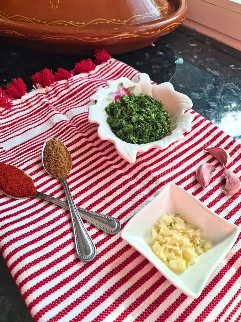 Prepped ingredients for authentic Moroccan Chermoula marinade, featuring a bowl of finely chopped fresh herbs, minced garlic, whole garlic cloves, and spoons holding bright paprika and ground cumin, arranged on a red and white striped cloth in front of a traditional clay tagine.