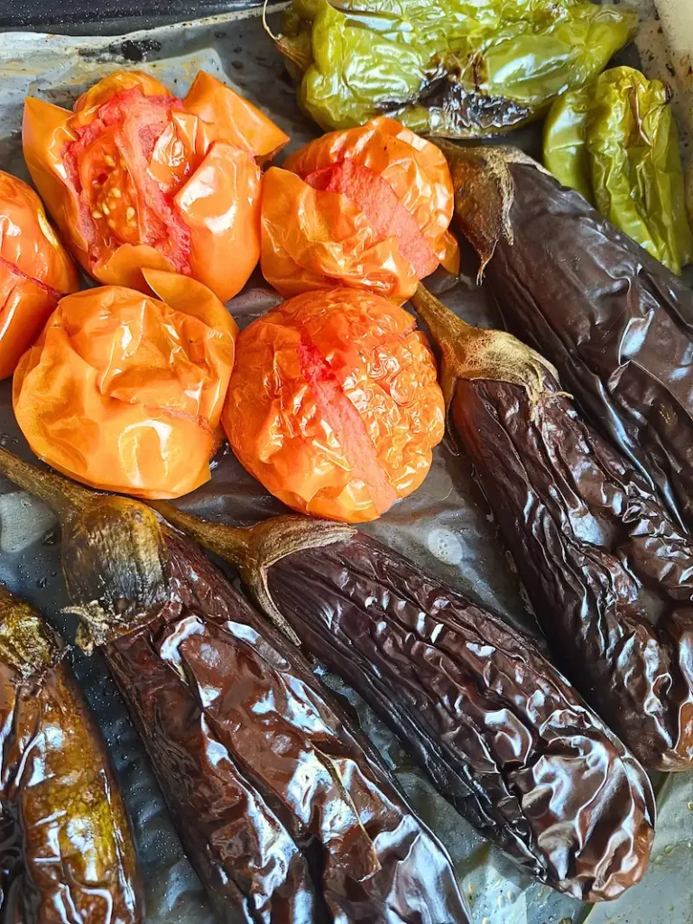 Freshly roasted eggplant, tomatoes, and a green pepper steaming on a metal baking tray, the essential first step for a smoky roasted eggplant and tomato salad.