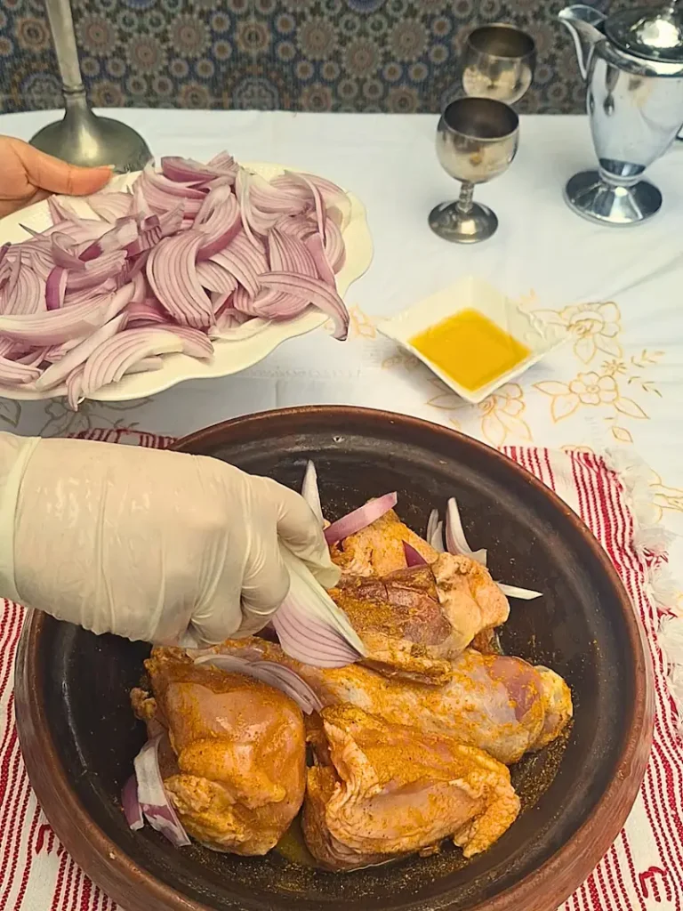 A step-by-step cooking photo showing raw, saffron-marinated chicken topped with a thick layer of freshly sliced raw onions inside a cooking pot, demonstrating how to prep a Chicken Tagine with Caramelized Onions.