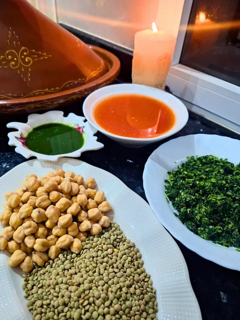 Prepped ingredients for authentic Moroccan Harira, featuring a plate of soaked chickpeas and green lentils, bowls of fresh blended tomato sauce and crushed herbs, with a traditional clay tagine in the background.