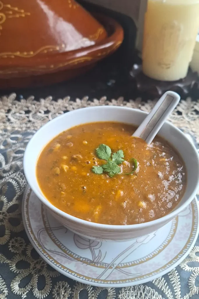 A Warm Bowl of Moroccan Ramadan Soup A steaming bowl of authentic Moroccan Harira soup filled with hearty chickpeas and lentils, garnished with fresh cilantro, resting on a lace tablecloth with a traditional clay tagine and a lit candle in the background.