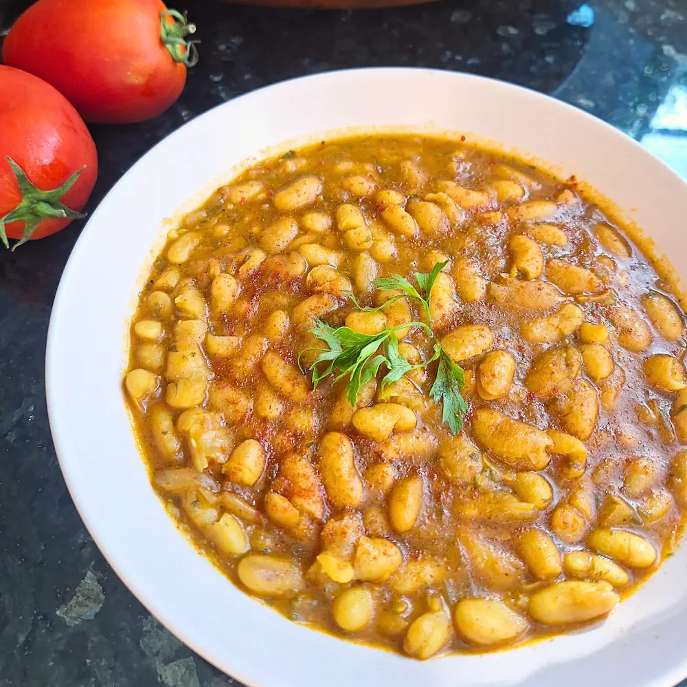 A top-down close-up of authentic Moroccan Loubia, showing tender, creamy white beans coated in a rich, warm tomato broth. The dish is garnished with smoked paprika and fresh parsley, sitting next to fresh red tomatoes.
