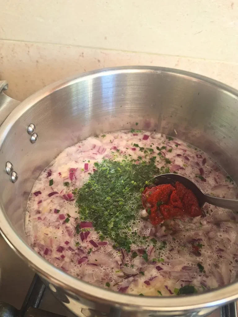 A stainless steel pressure cooker filled with water, soaked white beans, finely chopped red onions, crushed garlic, and fresh green herbs, with a spoon adding rich red tomato paste just before the spices are mixed in to make authentic Moroccan Loubia.