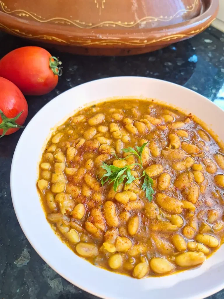 A vertical close-up of traditional Moroccan Loubia, a rich and velvety vegan white bean stew dusted with smoked paprika and garnished with fresh parsley. In the background sits a traditional clay tagine and fresh red tomatoes on a dark countertop.