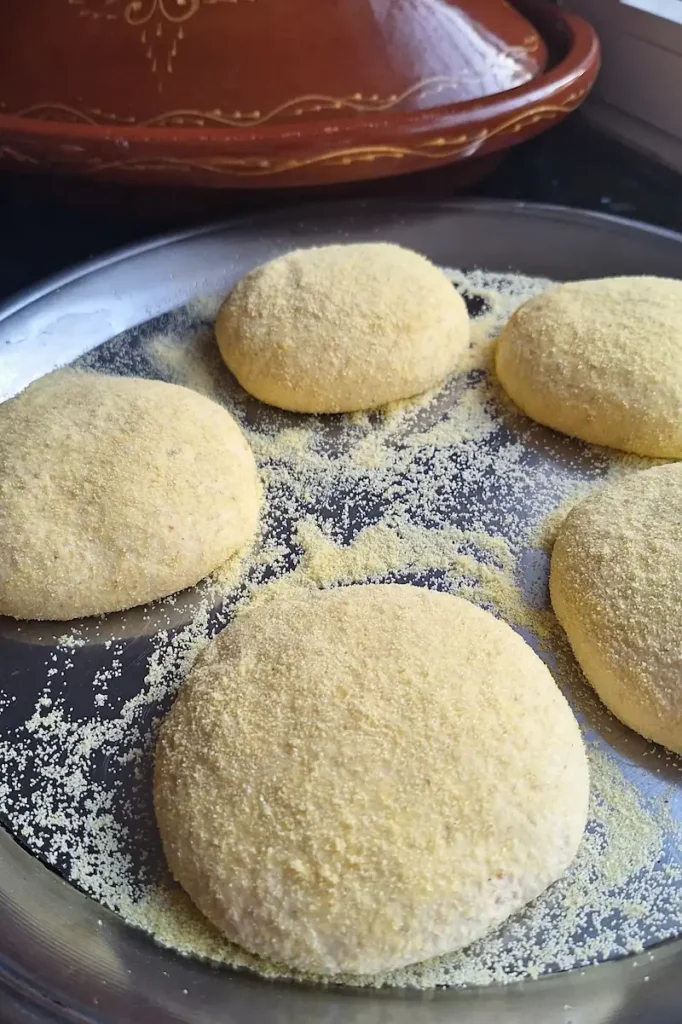 Five smooth, round balls of authentic Moroccan Batbout dough resting on a metal tray, generously dusted with extra-fine semolina flour before being flattened into pan bread. A traditional clay tagine sits blurred in the background.