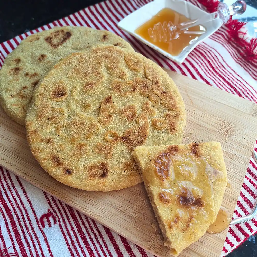 A square close-up of authentic Moroccan Batbout, a golden stovetop semolina pan bread, resting on a wooden board. A wedge is drizzled with glistening honey, with a small dish of honey and a traditional red-striped towel in the background.