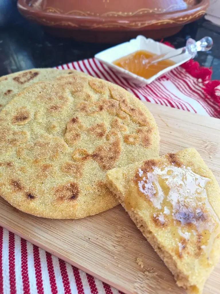 A vertical shot of traditional Moroccan Batbout (stovetop pan bread) resting on a wooden board with a honey-drizzled wedge. A red and white towel and a traditional clay tagine sit in the background.
